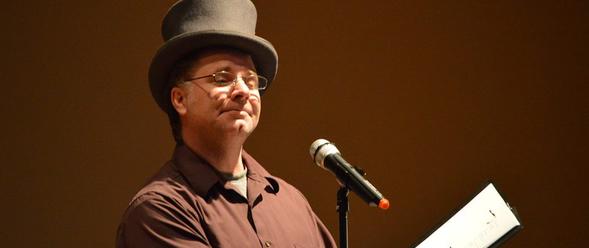 A photo of a man in a brown shirt and top hat standing in front of a microphone. 