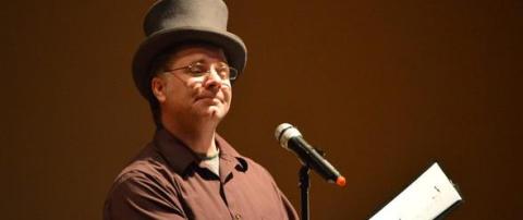 A photo of a man in a brown shirt and top hat standing in front of a microphone. 