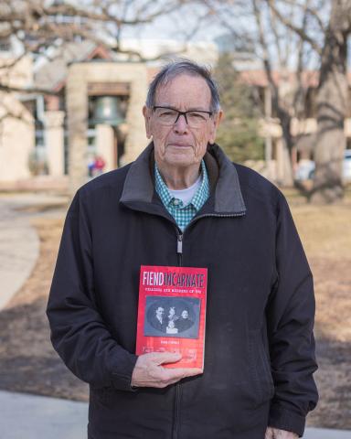 A photo of Ed Epperly (man with glasses, gray hair, and a brown coat), holding a copy of his book Fiend Incarnate. 