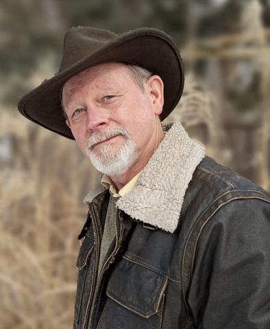 A photo of William Kent Krueger (a man with a brown cowboy hat, and white hair and beard, wearing a brown jacket) with a field of wild grasses behind him. 