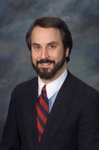 A photo of Jeff Stein, a man with brown hair and a brown beard. He is wearing a navy suit, white shirt, and a striped navy and red tie. 