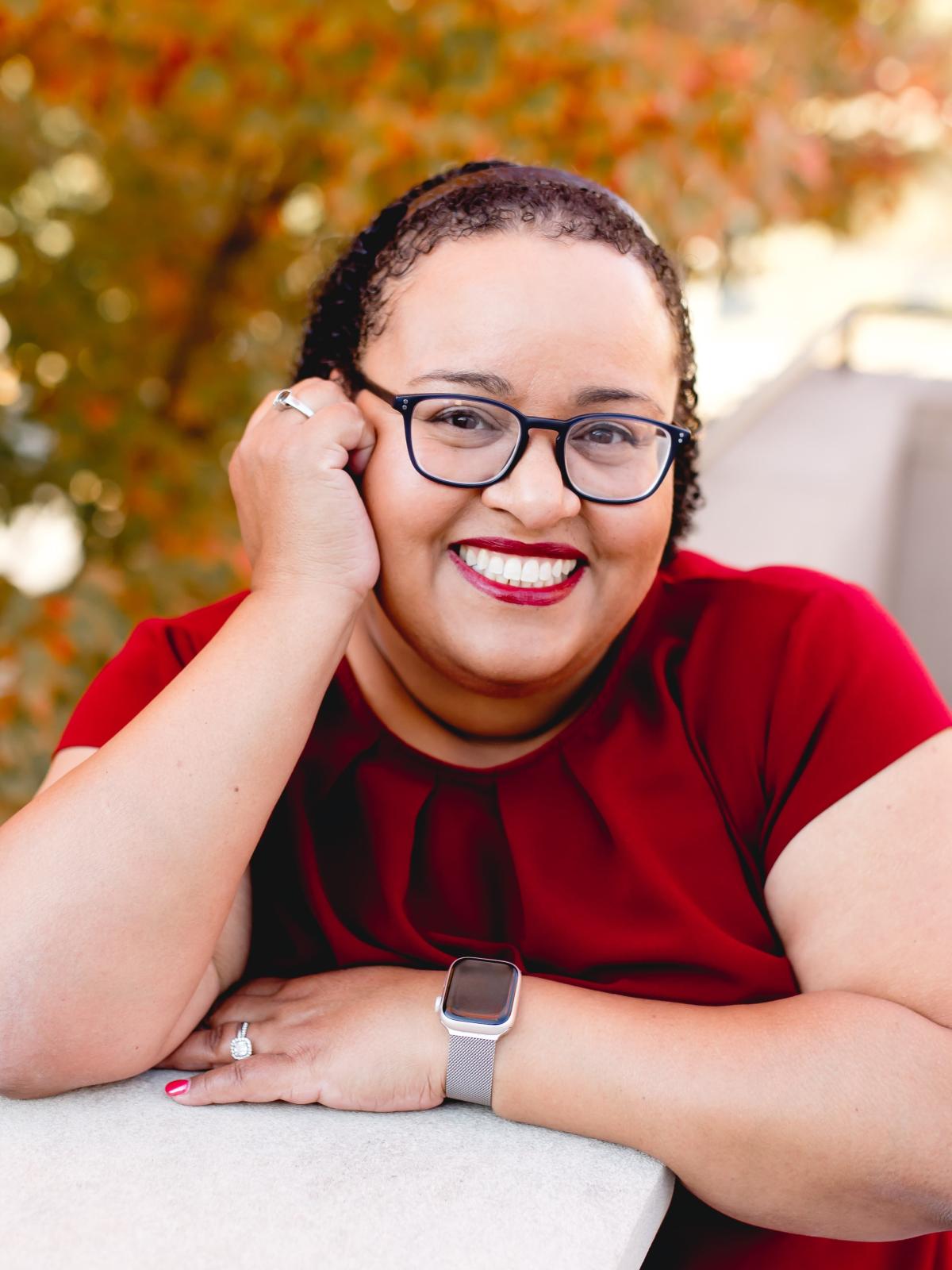 A photo of author Denise Williams, a woman with black curly hair, glasses, and a red shirt. 